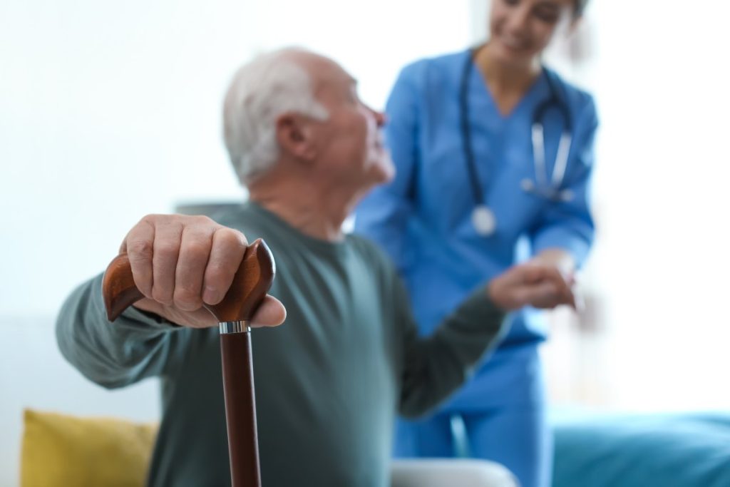 Anziano con bastone seduto e infermiera che lo aiuta ad alzarsi