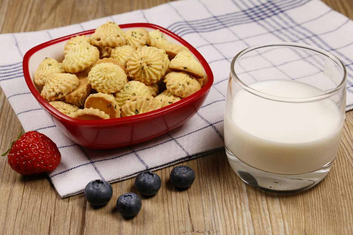 ciotola di biscotti accanto ad un bicchiere di latte un fragola e dei mirtilli