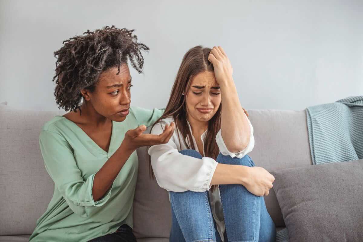 Donna di colore vestita di verde consola altra donna che piange con capelli lisci, seduta accanto a lei sul divano